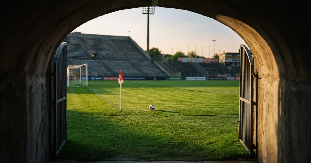 Blick durch ein Stadiontor auf einen grünen Rasen im Abendlicht, ein Ball liegt an der Eckfahne, die Ränge sind leer.