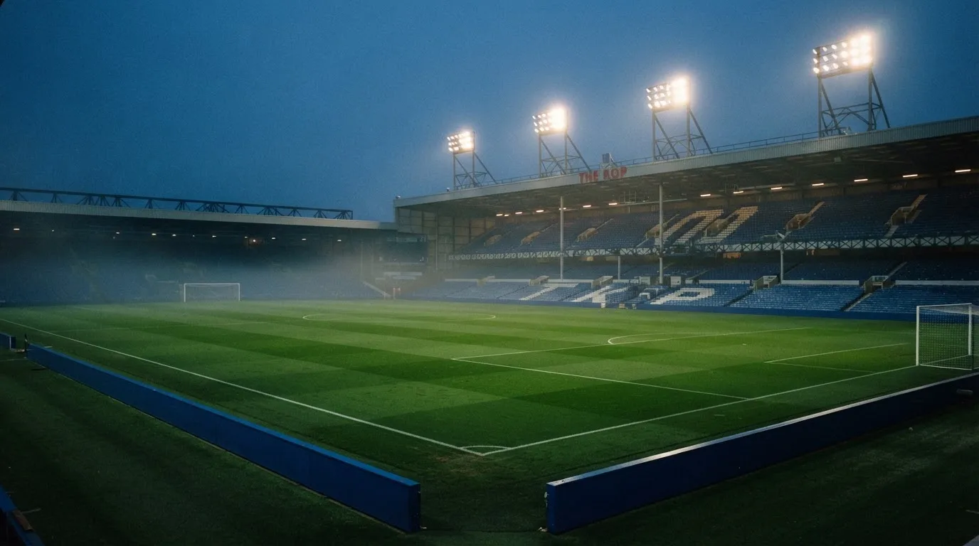 Eine leere Stadiontribüne unter Flutlicht aus der Ferne betrachtet, die Rasenfläche leuchtet grün in der Dämmerung.