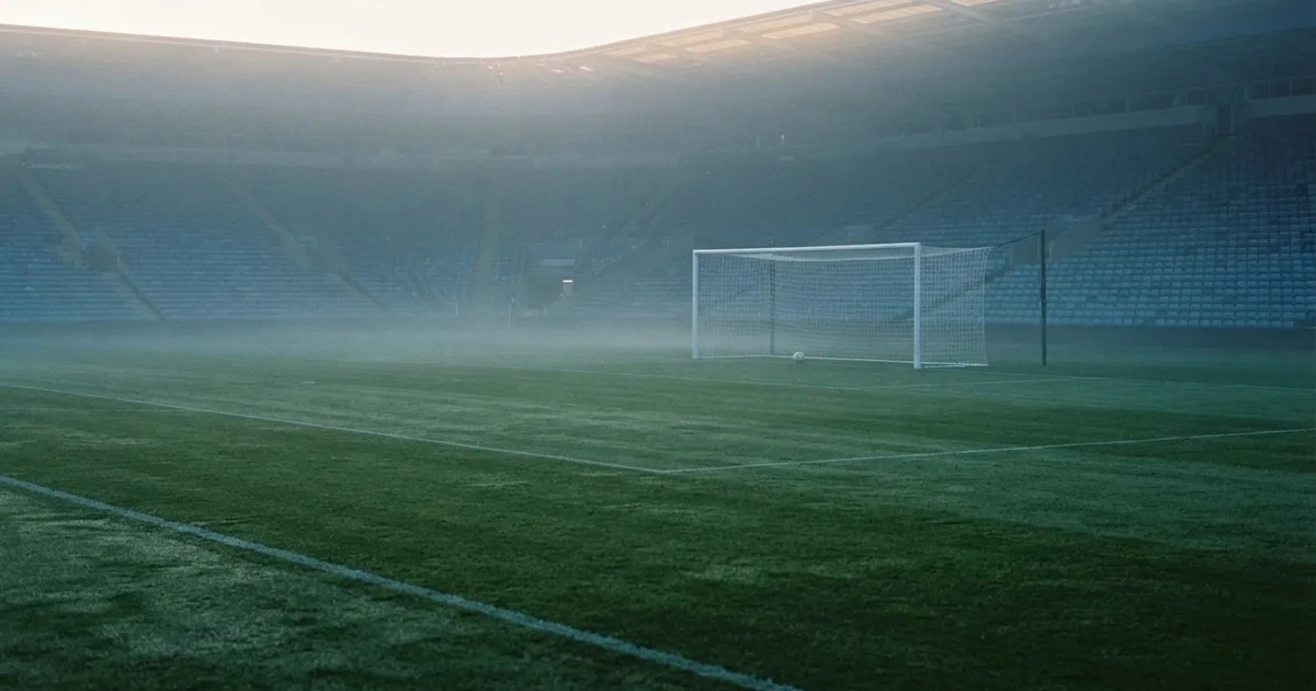 Ein Fußballplatz mit gepflegtem Rasen und einem Tor, über dem Spielfeld liegt Morgennebel, im Hintergrund eine leere Tribüne.