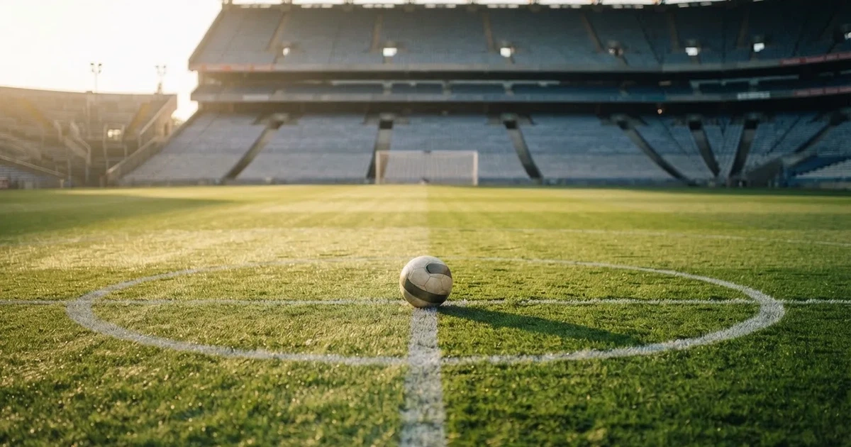 Ein Fußball liegt auf dem Mittelkreis eines Stadionrasens am frühen Morgen, im Hintergrund leere Ränge ohne erkennbare Beschriftung.
