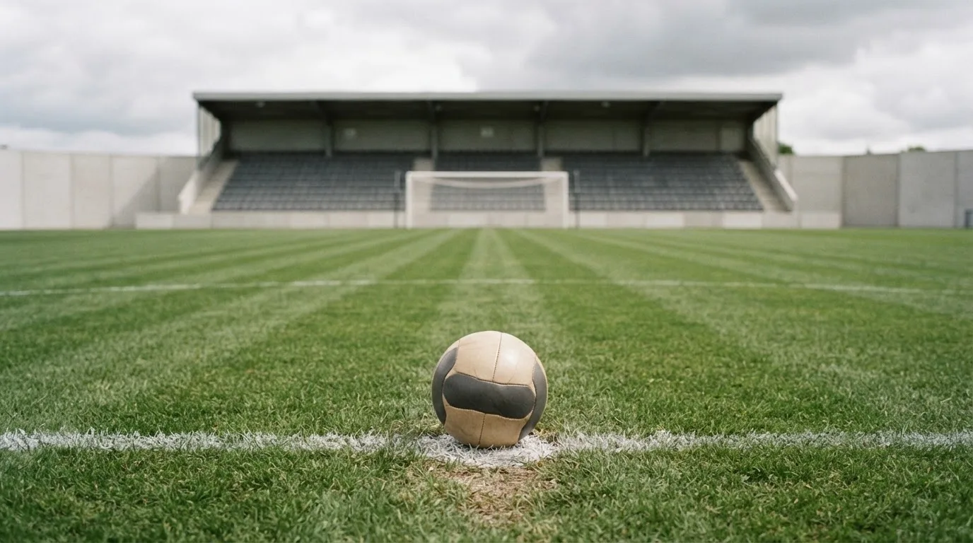 Ein Fußball liegt auf dem Anstoßpunkt eines leeren Rasenplatzes, im Hintergrund eine ruhige Tribüne unter bedecktem Himmel.