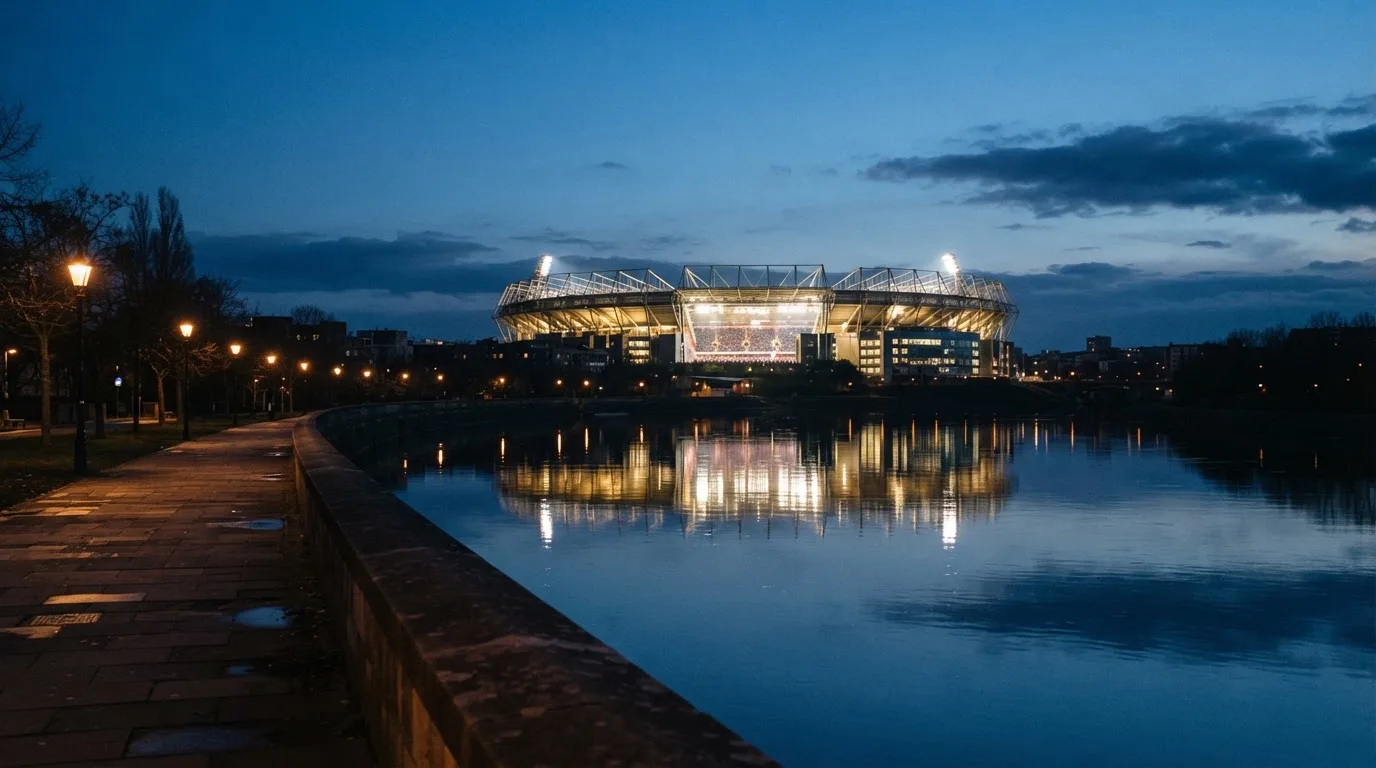 Ein Stadion unter Flutlicht aus der Ferne gesehen, im Vordergrund ein ruhiger Fluss und ein Fußweg.