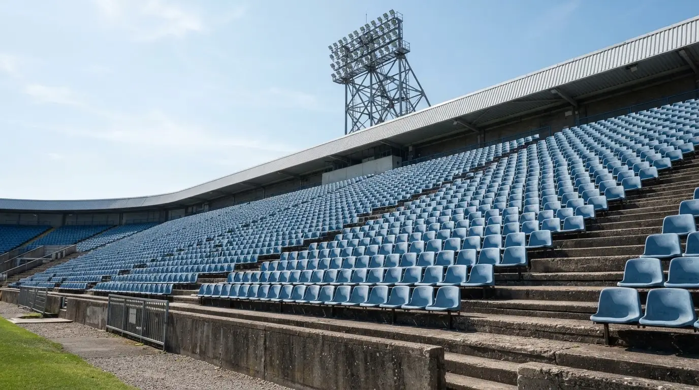 Eine leere Tribüne eines Fußballstadions bei Tageslicht, Sitzschalen in Reihen und dahinter ein Flutlichtmast.