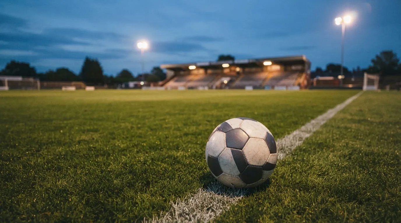 Ein Fußball liegt auf dem Rasen eines leeren Platzes, im Hintergrund ein Stadion unter Flutlicht in der Abenddämmerung.