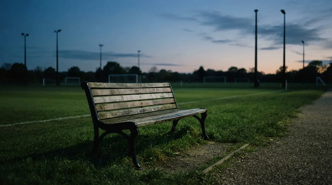 Eine leere Parkbank am Rand eines Fußballfelds in der Abenddämmerung, im Hintergrund ragen dunkle Flutlichtmasten in den Himmel.