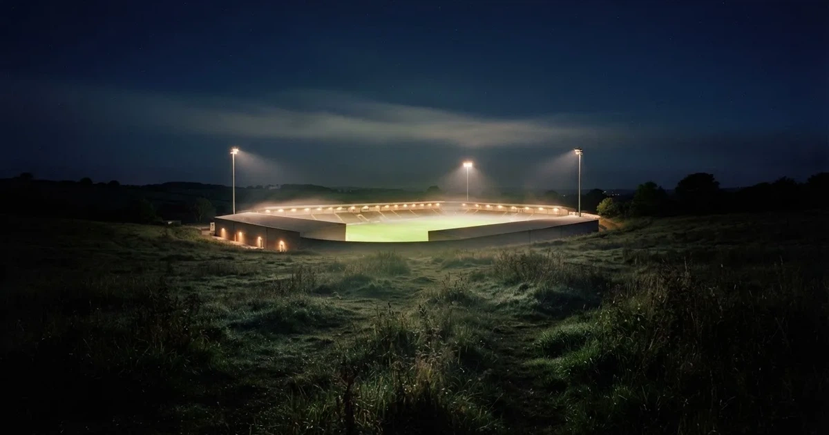 Ein Stadion unter Flutlicht, aus großer Entfernung über eine dunkle Wiese fotografiert, am Himmel zieht Nebel auf.