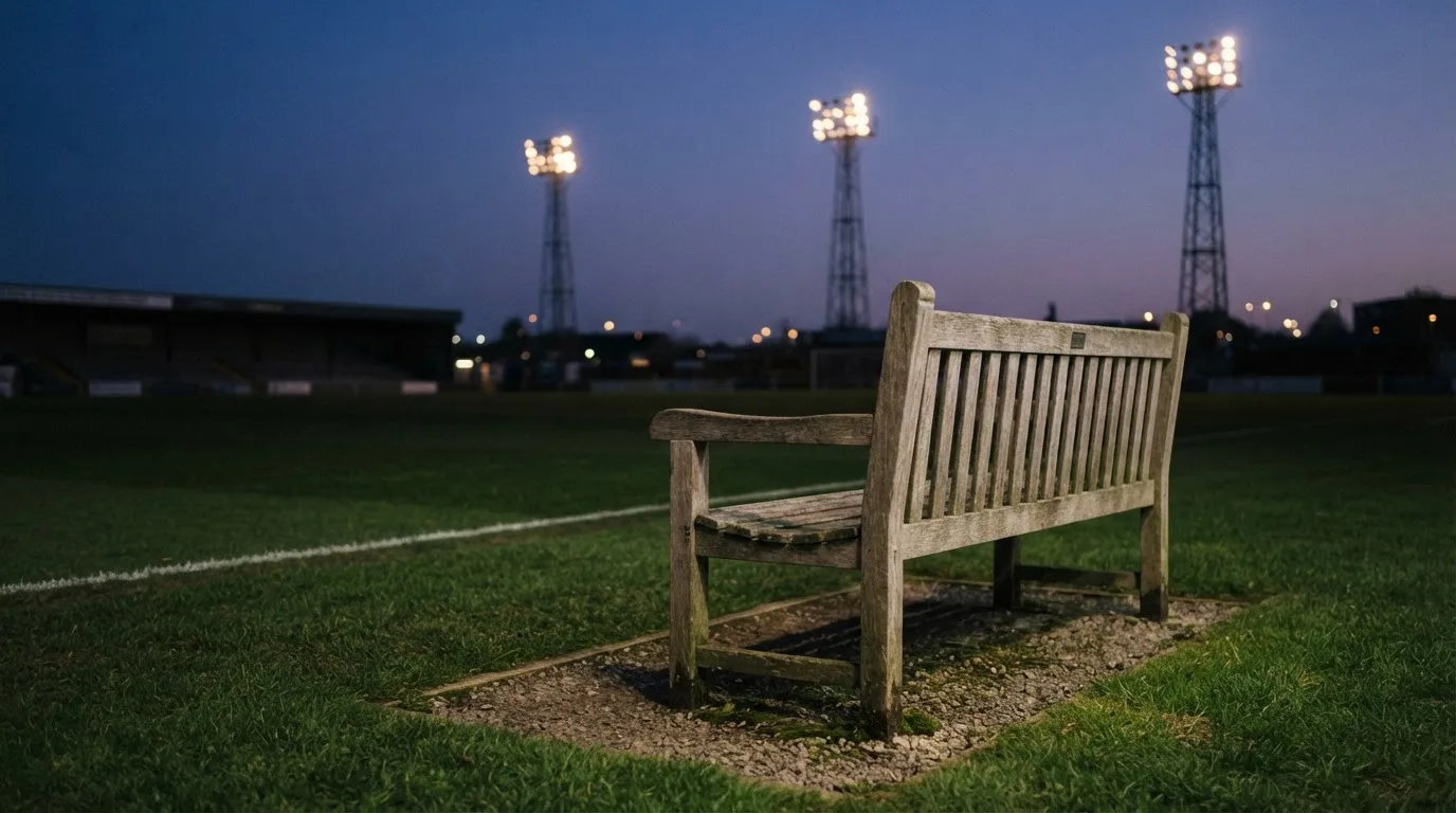 Eine leere Parkbank am Rand eines Fußballfelds in der Abenddämmerung, im Hintergrund ragen dunkle Flutlichtmasten in den Himmel.