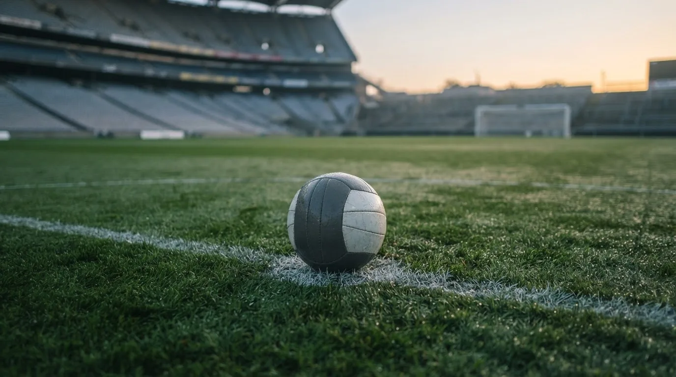 Ein Fußball liegt auf dem Anstoßpunkt eines leeren Rasenplatzes in der Morgendämmerung, im Hintergrund eine leere Tribüne.