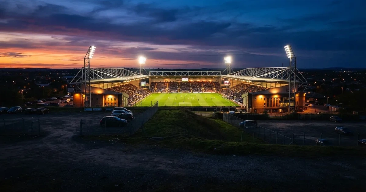 Ein Fußballstadion unter Flutlicht aus großer Entfernung, davor ein dunkler Parkplatz und ein heller Abendhimmel.