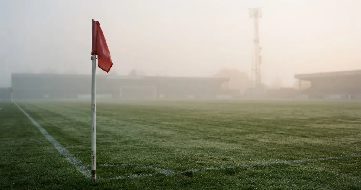 Eine Eckfahne steht im Nebel am Rand eines leeren Rasenplatzes in der Morgendämmerung, dahinter ein Flutlichtmast.