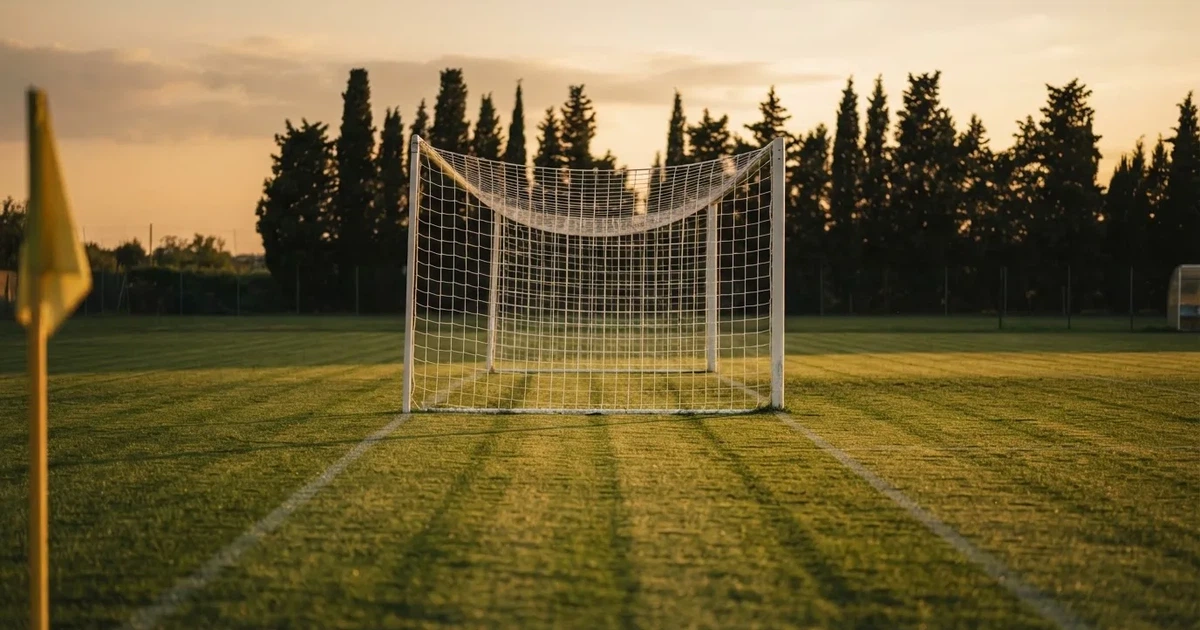 Ein Fußballtor mit Netz auf einem leeren Platz im Abendlicht, der Rasen frisch gemäht, im Hintergrund Zypressen.