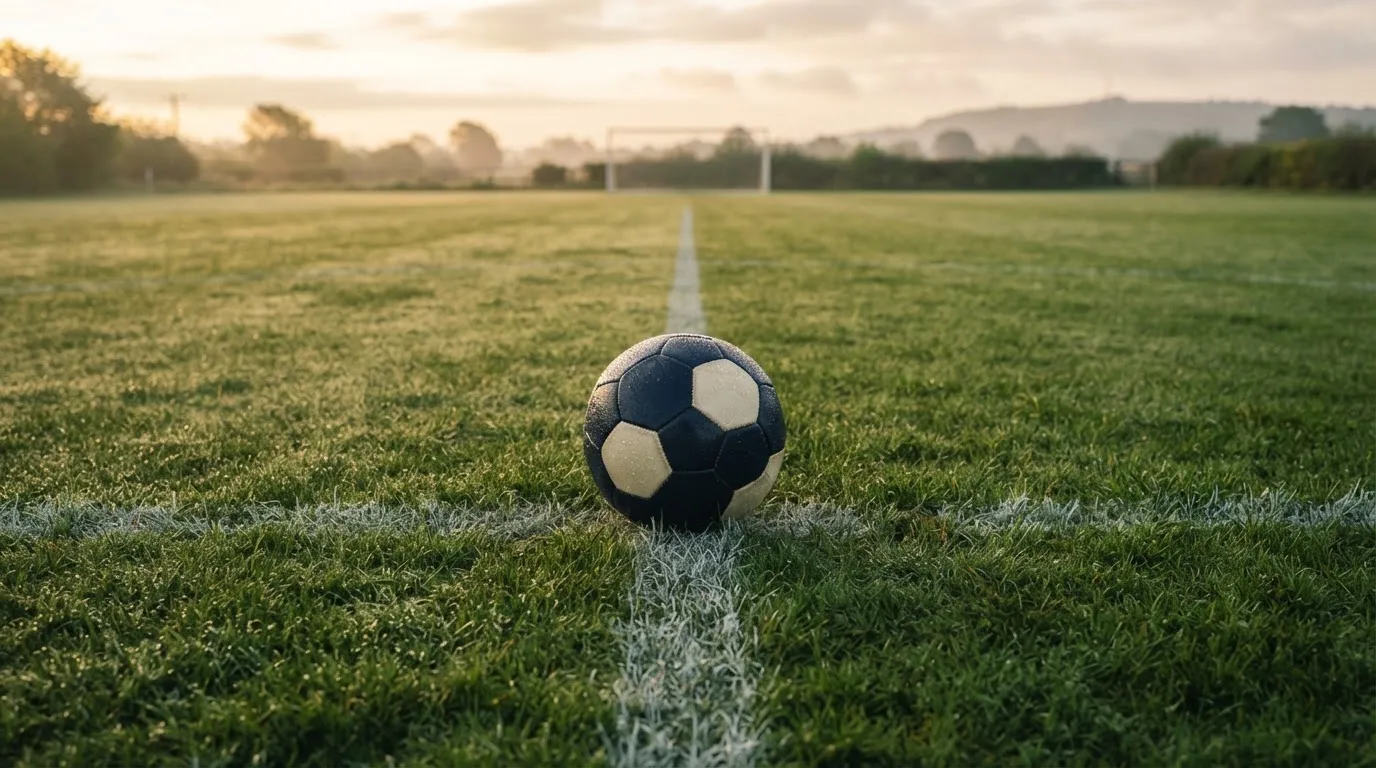 Ein Fußball ruht auf dem Mittelkreis eines leeren Rasenplatzes in der Morgendämmerung, Tau liegt auf dem Gras.