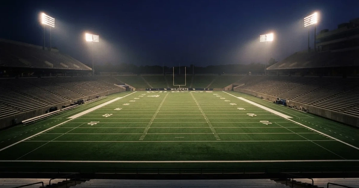 Ein American-Football-Feld mit weißen Yardlinien im Flutlicht, von einer hohen Tribüne aus gesehen, die Ränge leer, Zahlen nicht erkennbar.