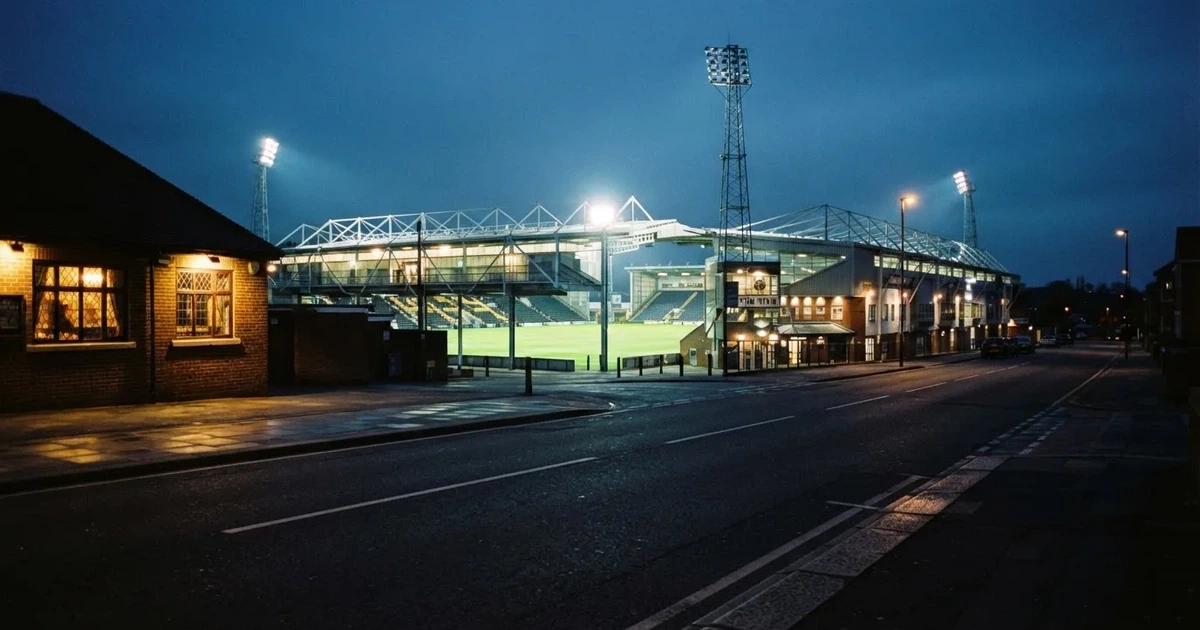 Ein Fußballstadion bei Flutlicht aus der Ferne gesehen, davor eine Straße und leuchtende Fenster eines Nebengebäudes.