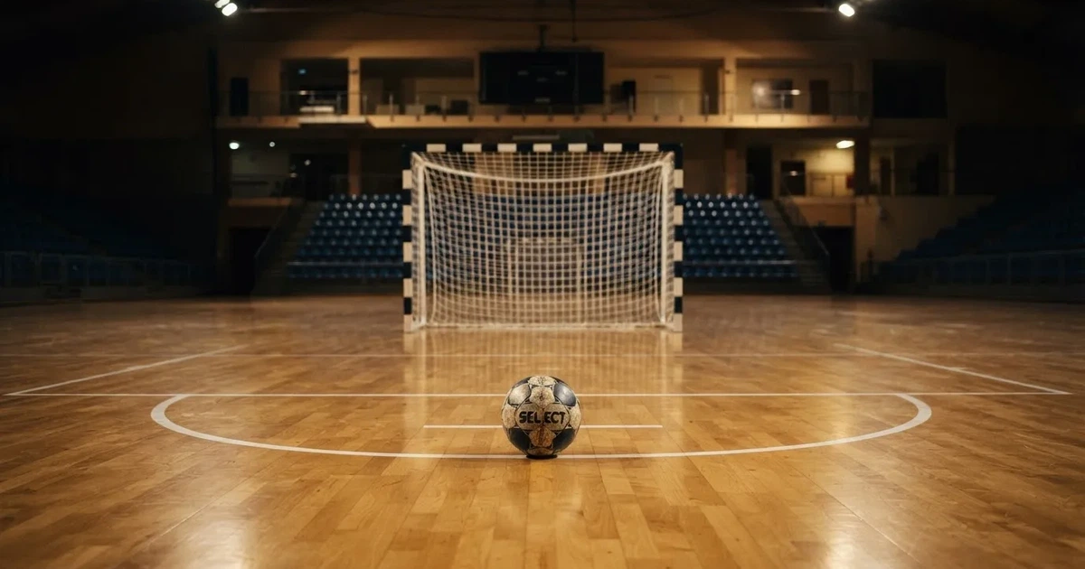 Ein Handball liegt auf dem Hallenboden vor einem Tor mit Netz, die leere Halle wird von Flutlicht beleuchtet.