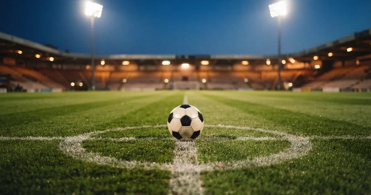 Ein Rasenplatz in der Dämmerung mit eingeschalteten Flutlichtmasten, ein Ball liegt am Anstoßpunkt, die Tribünen im Hintergrund sind unscharf.