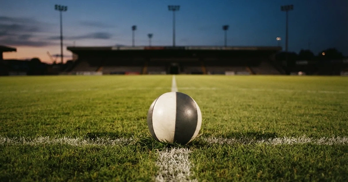 Ein Fußball liegt auf dem Anstoßpunkt eines leeren Rasenplatzes, im Hintergrund eine Tribüne und Flutlichtmasten in der Abenddämmerung.