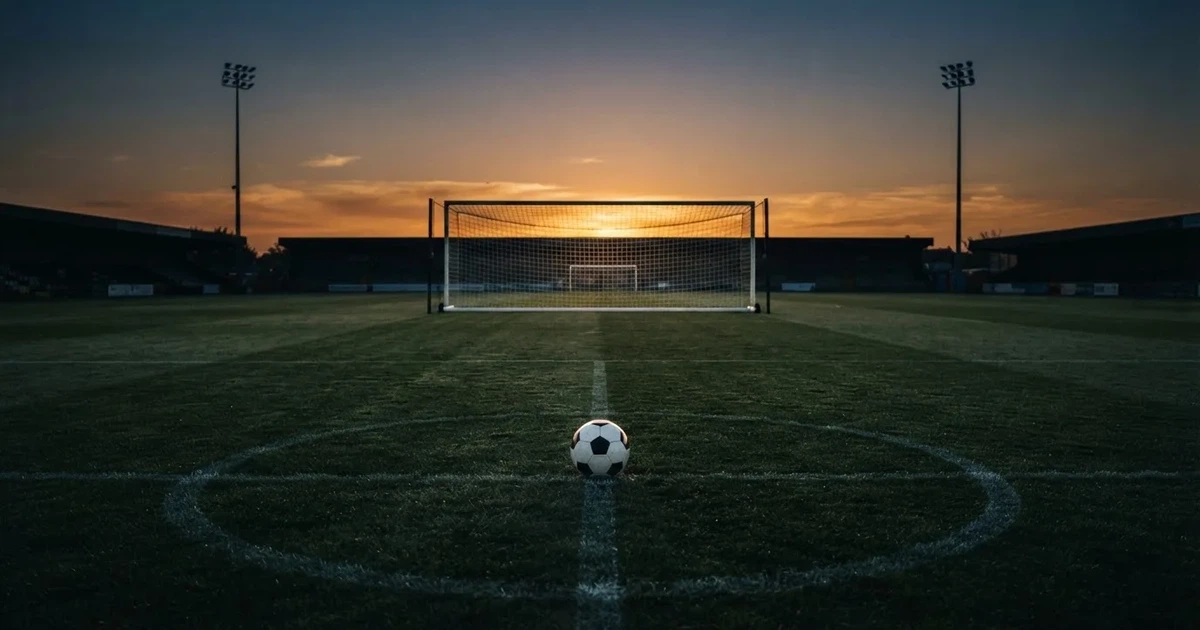 Ein Fußballtor mit Netz im Gegenlicht auf einem leeren Platz am Abend, davor ein Ball auf dem Rasen.