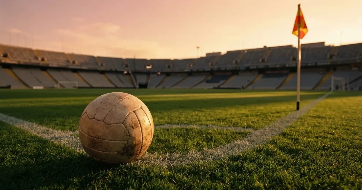 Ein Fußball liegt am Viertelkreis an der Eckfahne eines Rasenplatzes, im Hintergrund leere Tribünen in warmem Abendlicht.