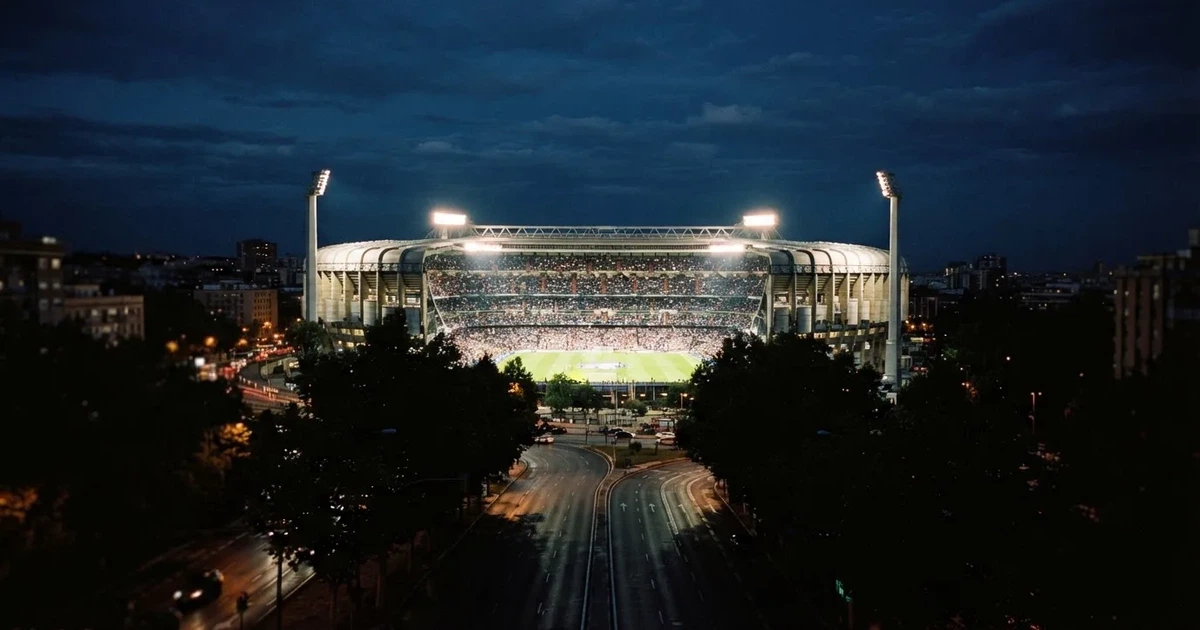 Ein Fußballstadion bei Nacht aus großer Entfernung, hell erleuchtet unter Flutlicht, davor eine dunkle Straße und Bäume.