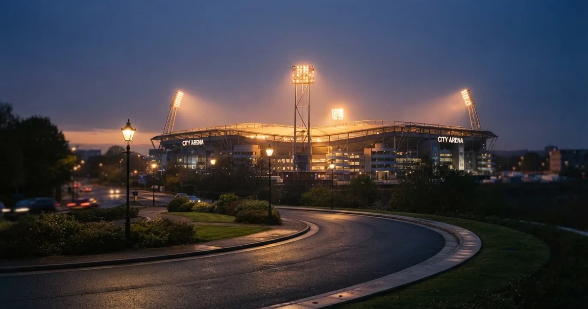 Ein Fußballstadion in der Dämmerung aus der Ferne, die Flutlichter gerade eingeschaltet, davor ein Kreisverkehr mit Laternen.